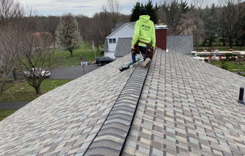 Technician in yellow hoodie, installing shingles on a gray roof. Trees in background.