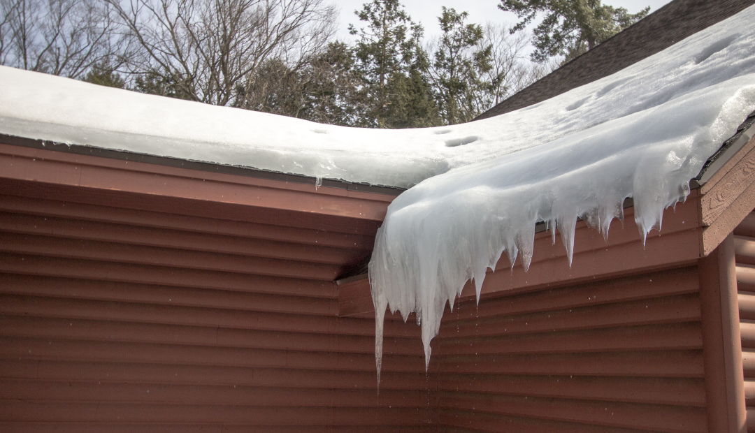 Ice dam and icicles on the edge of a snow-covered roof of a brown home.